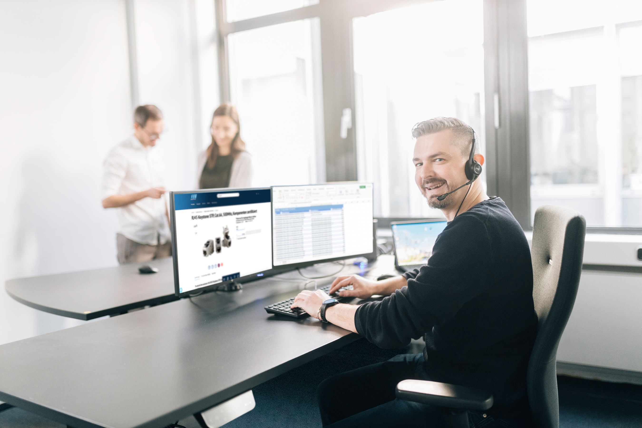 Mitarbeiter mit Headset am Schreibtisch mit zwei Monitoren, im Hintergrund zwei Personen im Gespräch