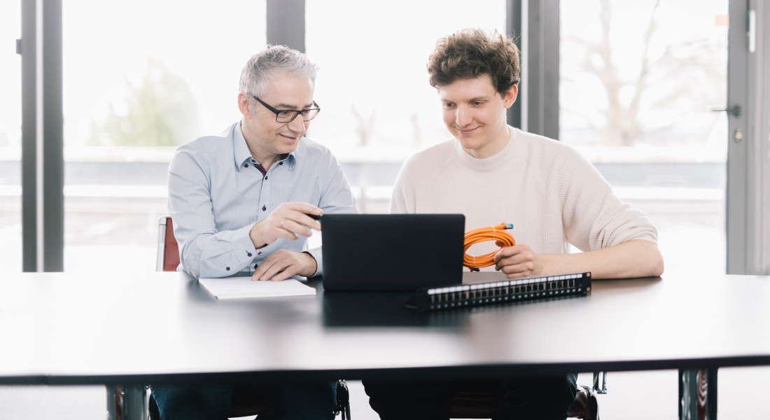 In einem Meeting an einem Tisch wird ein Kunde von EFB-Elektronik beraten. Auf dem Tisch steht ein Laptop. Der Berater hält ein Kabel in der Hand.