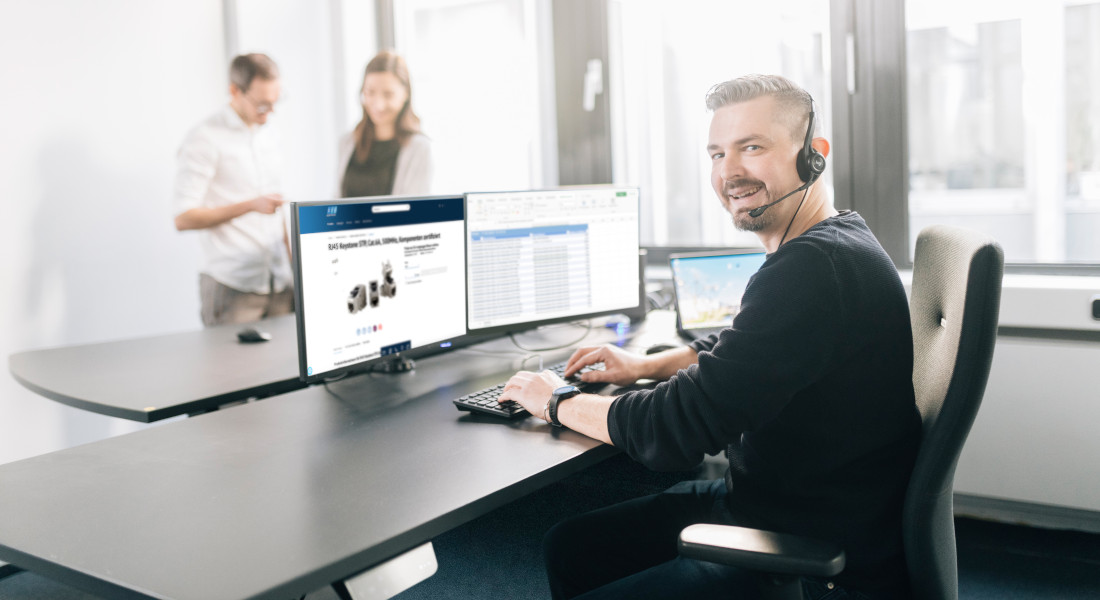 Mitarbeiter mit Headset am Schreibtisch mit zwei Monitoren, im Hintergrund zwei Personen im Gespräch